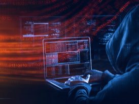 Person mit Kapuze sitzt vor Laptop und übt einen Cyberangriff aus
