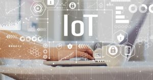 Eine Person tippt auf einem Laptop, darüber der Schriftzug „IoT“ sowie verschiedene Symbole zum Thema Cybersicherheit