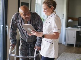 Senior Man In Dressing Gown Using Walking Frame Being Helped By Female Nurse With Digital Tablet