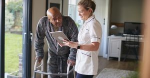 Senior Man In Dressing Gown Using Walking Frame Being Helped By Female Nurse With Digital Tablet