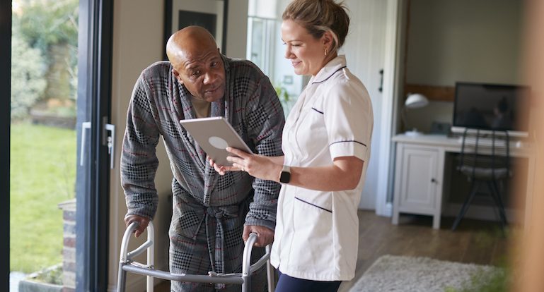 Senior Man In Dressing Gown Using Walking Frame Being Helped By Female Nurse With Digital Tablet