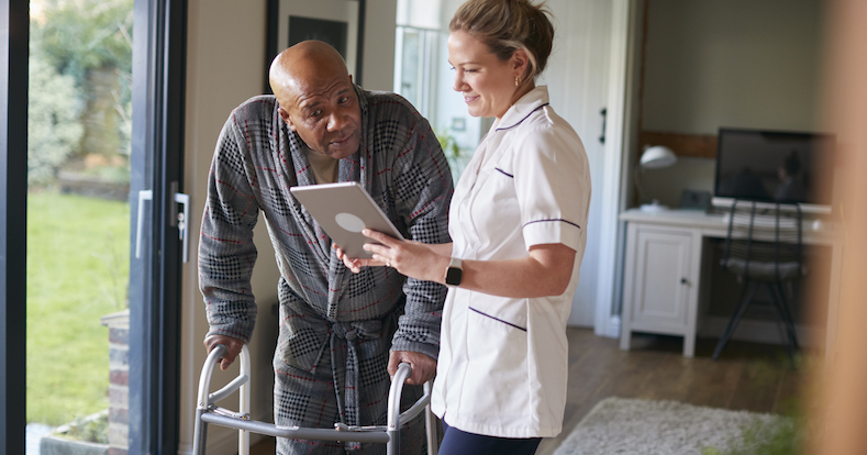 Senior Man In Dressing Gown Using Walking Frame Being Helped By Female Nurse With Digital Tablet